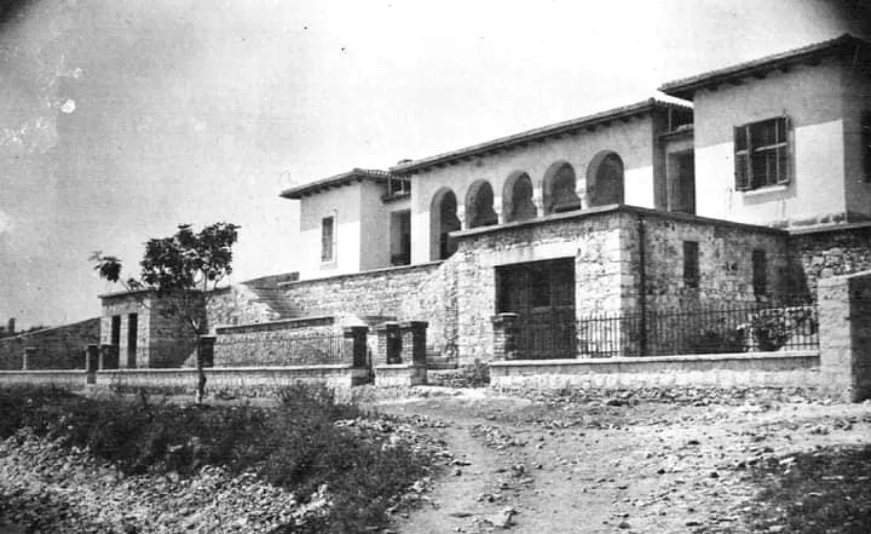 The front of a white two-storey house with four arches on the upper floor and a stone wall below, seen from a gravel track under a bright sky.