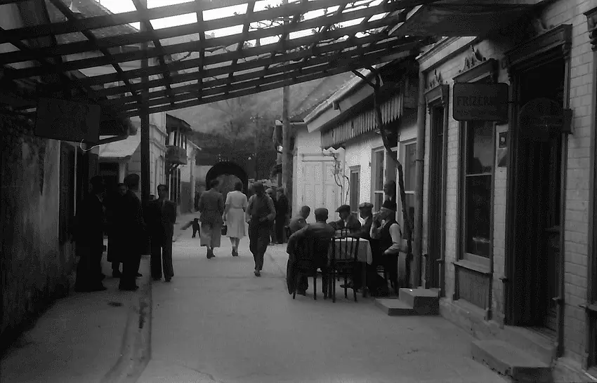 Fotografie alb-negru: o ulicioară cu pergolă de lemn, mese de cafenea, bărbați așezați, firme deasupra prăvăliilor.