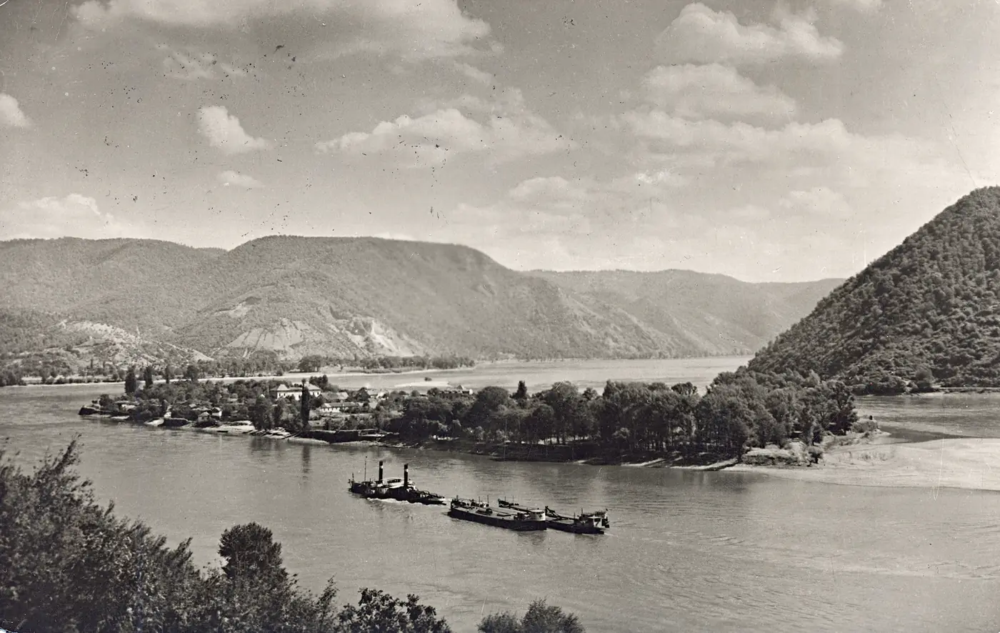 Fotografie alb-negru: insula Ada Kaleh, împădurită, în mijlocul Dunării, cu munții sârbești în spate și un remorcher cu șlepuri în prim-plan.