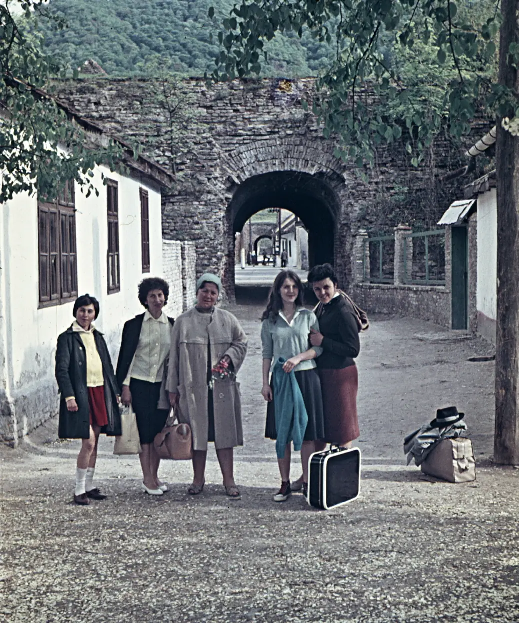 Fotografie color: un tunel de piatră prin zidul cetății, case albe de o parte și de alta, câteva turiste cu valize.