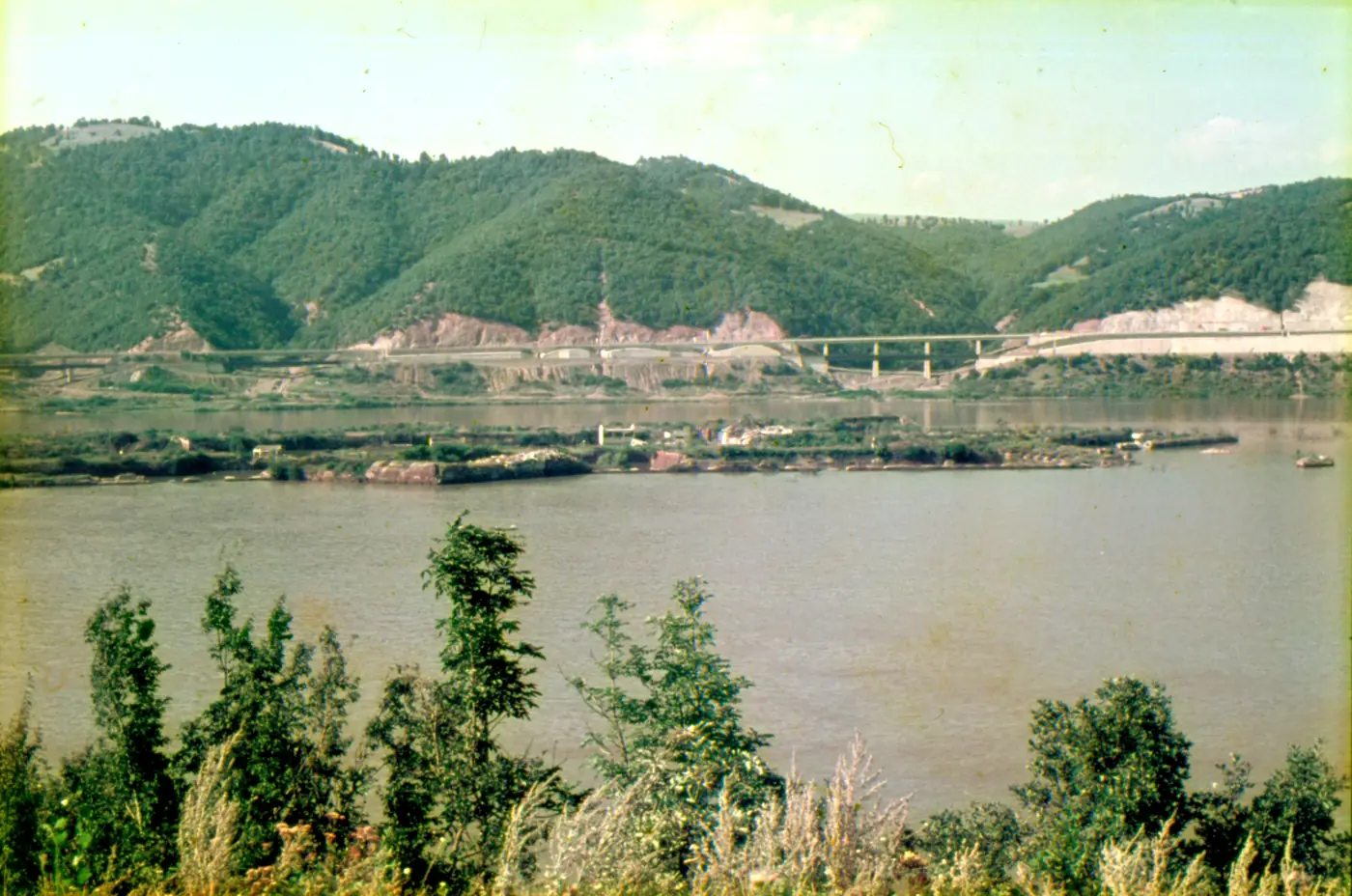 Fotografie color de pe malul sârbesc: insula rasă, abia deasupra apei, cu resturi de ziduri de cărămidă; pe malul românesc, șoseaua nouă și un viaduct.