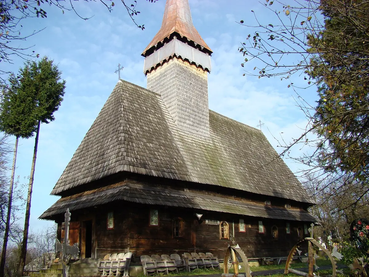 Biserica de lemn din Hoteni, cu acoperiș de șindrilă în două ape și turn zvelt, văzută de jos pe cer senin.