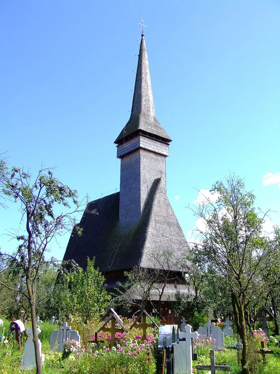 Biserica de lemn din Ieud, cu acoperiș abrupt de șindrilă și turn înalt terminat cu o fleșă ascuțită, într-un cimitir cu cruci.