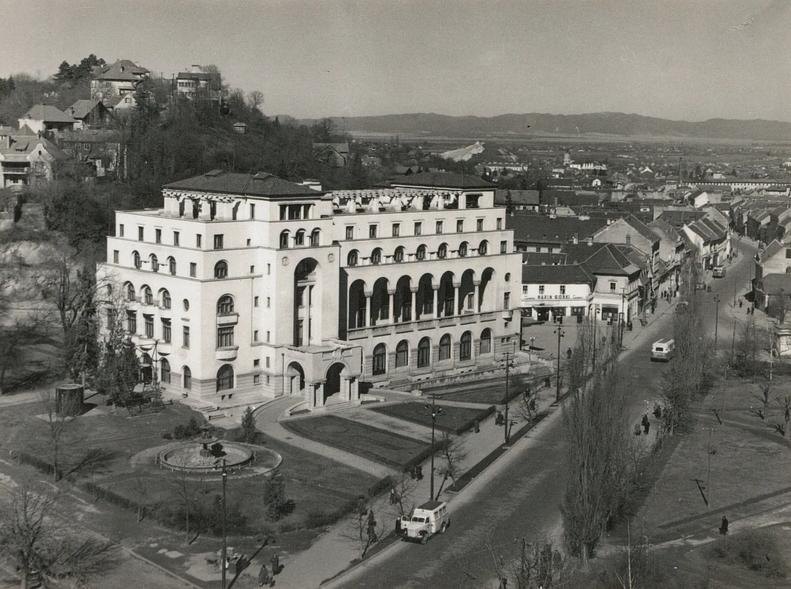 Fotografie alb-negru: clădire albă cu arcade, văzută de sus, cu bulevard și dealuri în spate