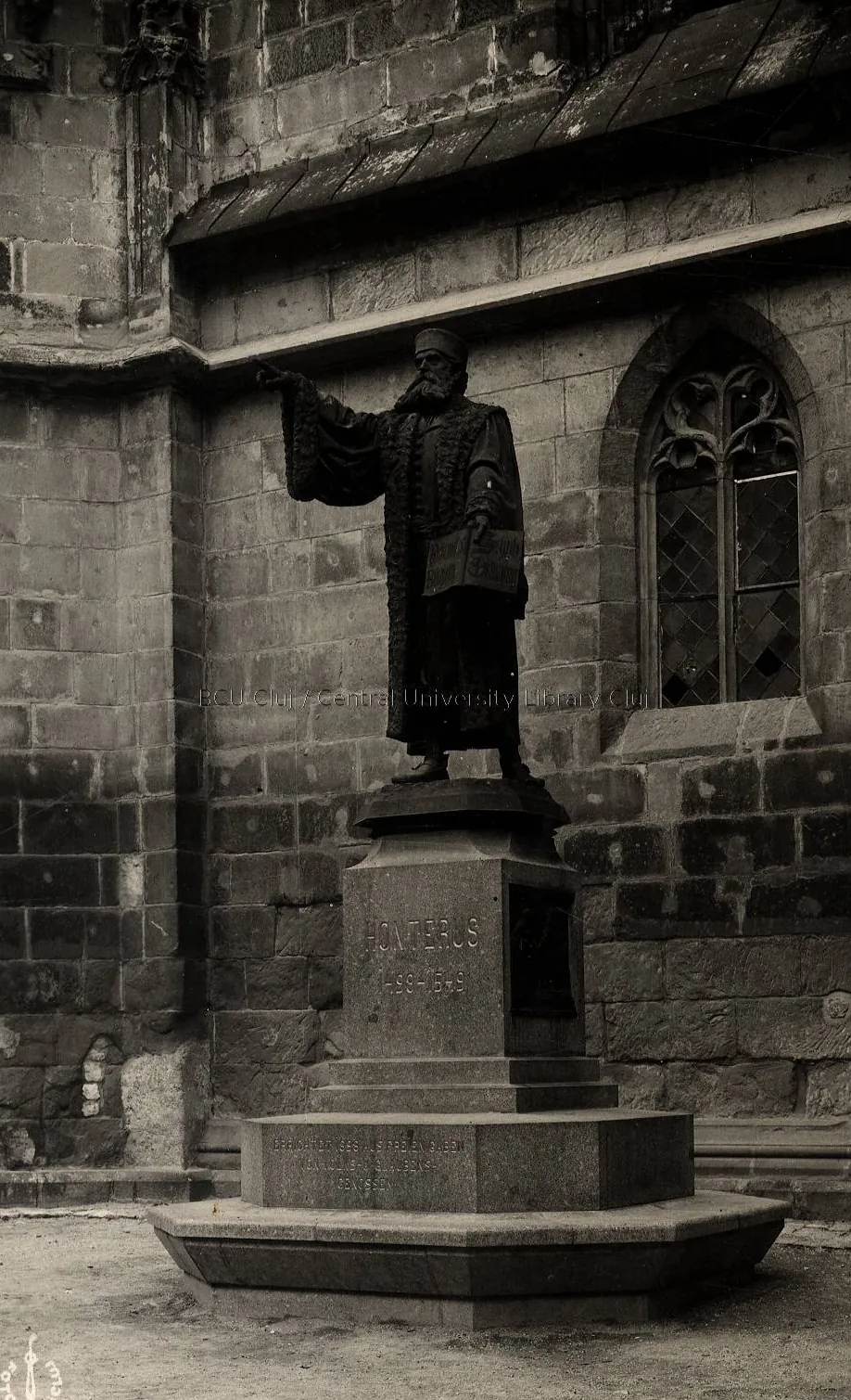 Fotografie alb-negru: statuia de bronz a lui Honterus lângă zidul gotic al Bisericii Negre