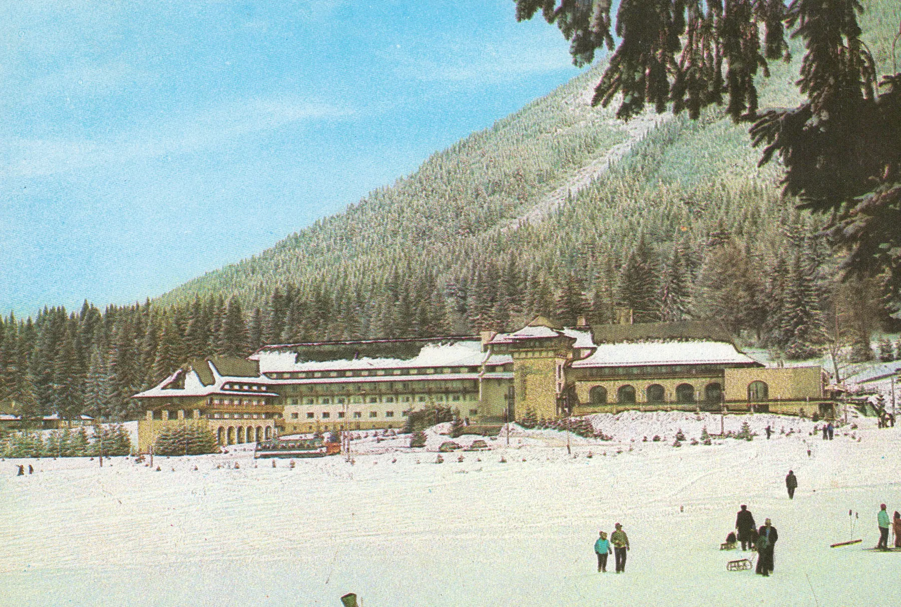 Colour postcard: a long hotel beneath a snowy slope, with tourists on the snow