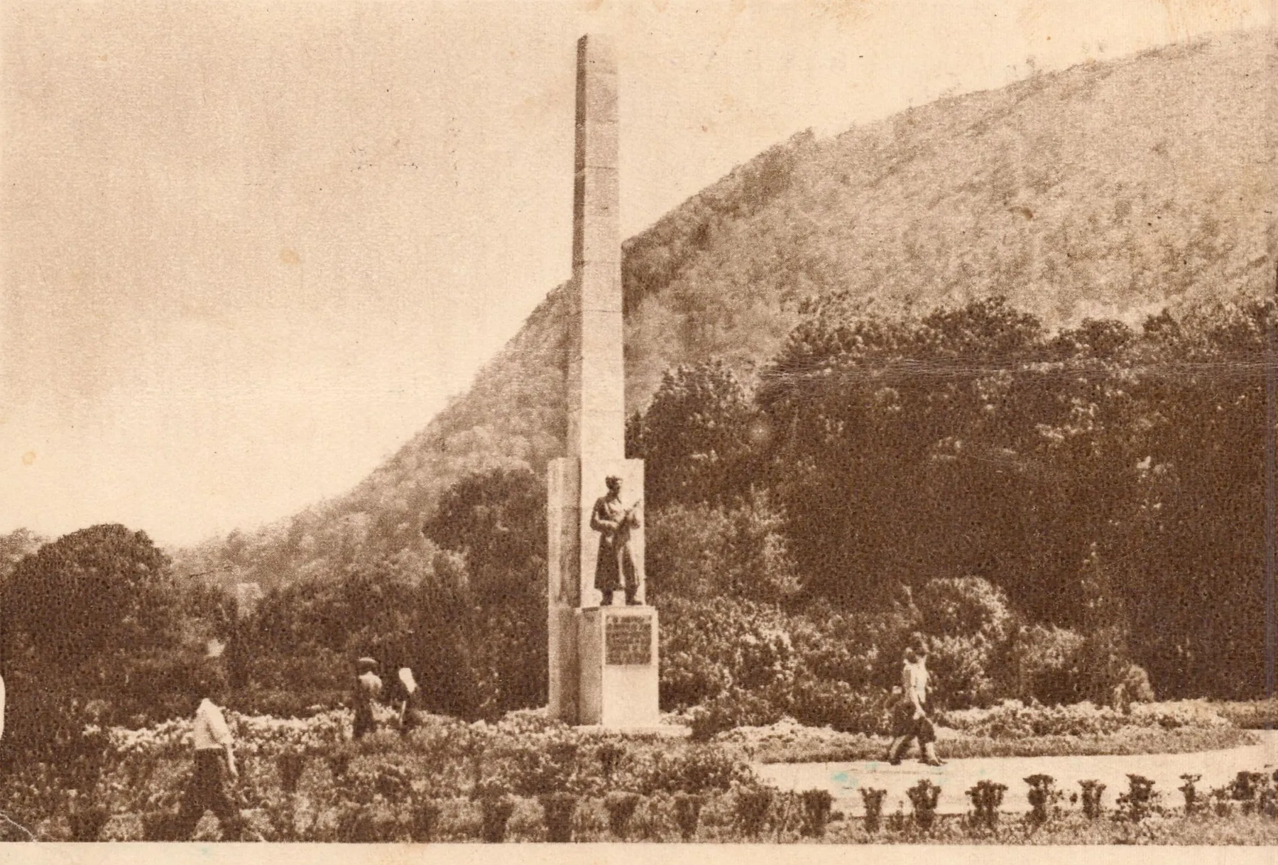 Carte poștală sepia: un obelisc înalt cu o statuie de soldat la bază, sub dealul împădurit