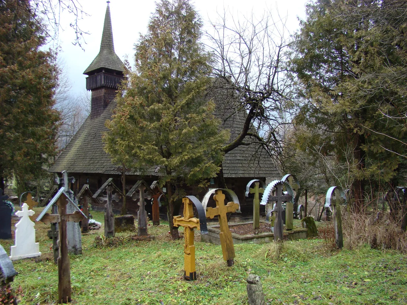 Biserica de lemn din Breb, cu turn înalt și acoperiș de șindrilă, văzută din cimitirul cu cruci de lemn și de metal.