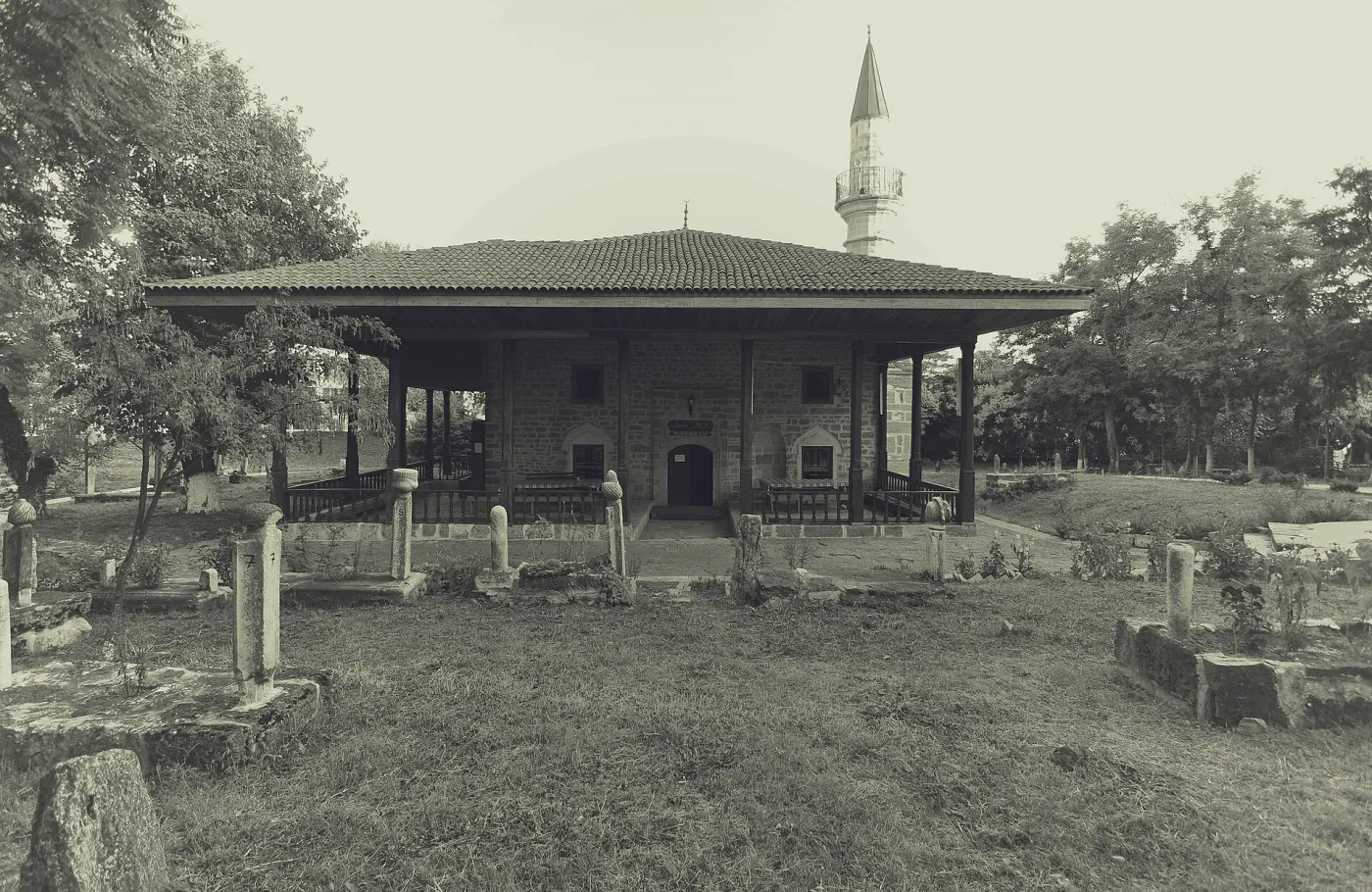 Geamia Esmahan Sultan din Mangalia, o clădire joasă de piatră cu pridvor pe stâlpi și un minaret subțire, înconjurată de pietre funerare vechi.