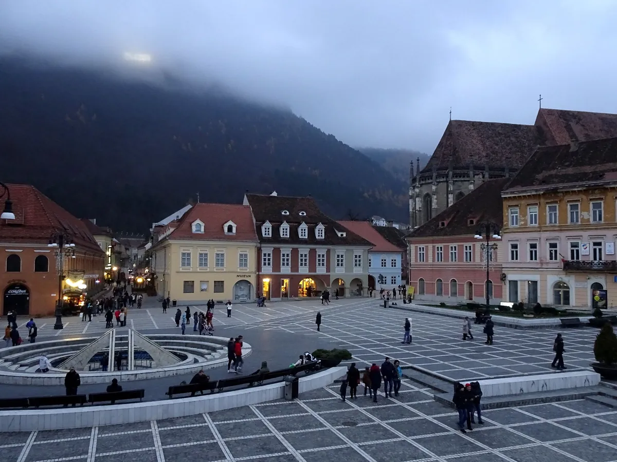 Piața Sfatului din Brașov seara, cu oameni, o fântână, case cu acoperișuri roșii și un munte în ceață.