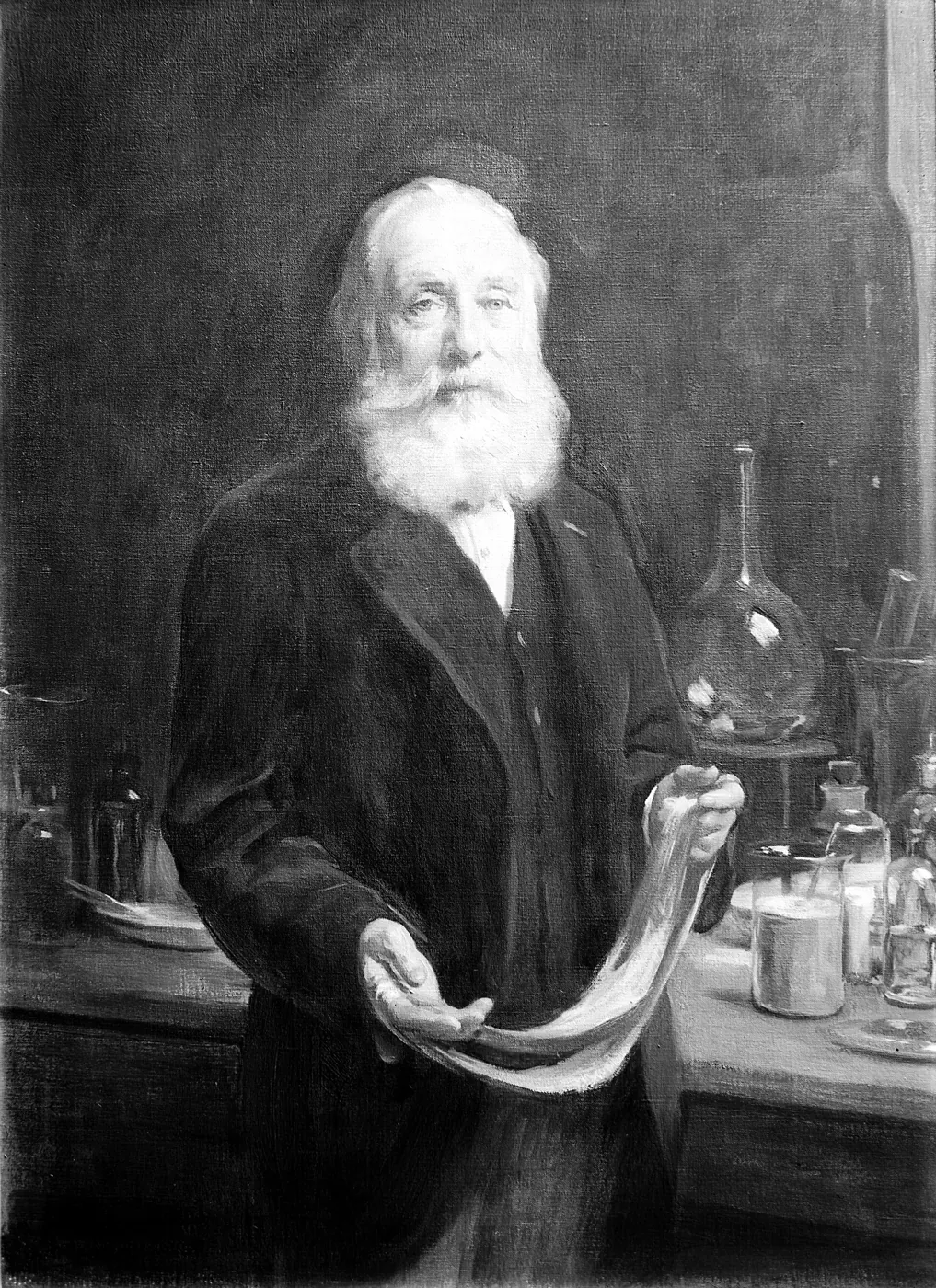 Portret alb-negru al unui bătrân cu barbă albă, în haină închisă la culoare, ținând o bucată de stofă într-un laborator.