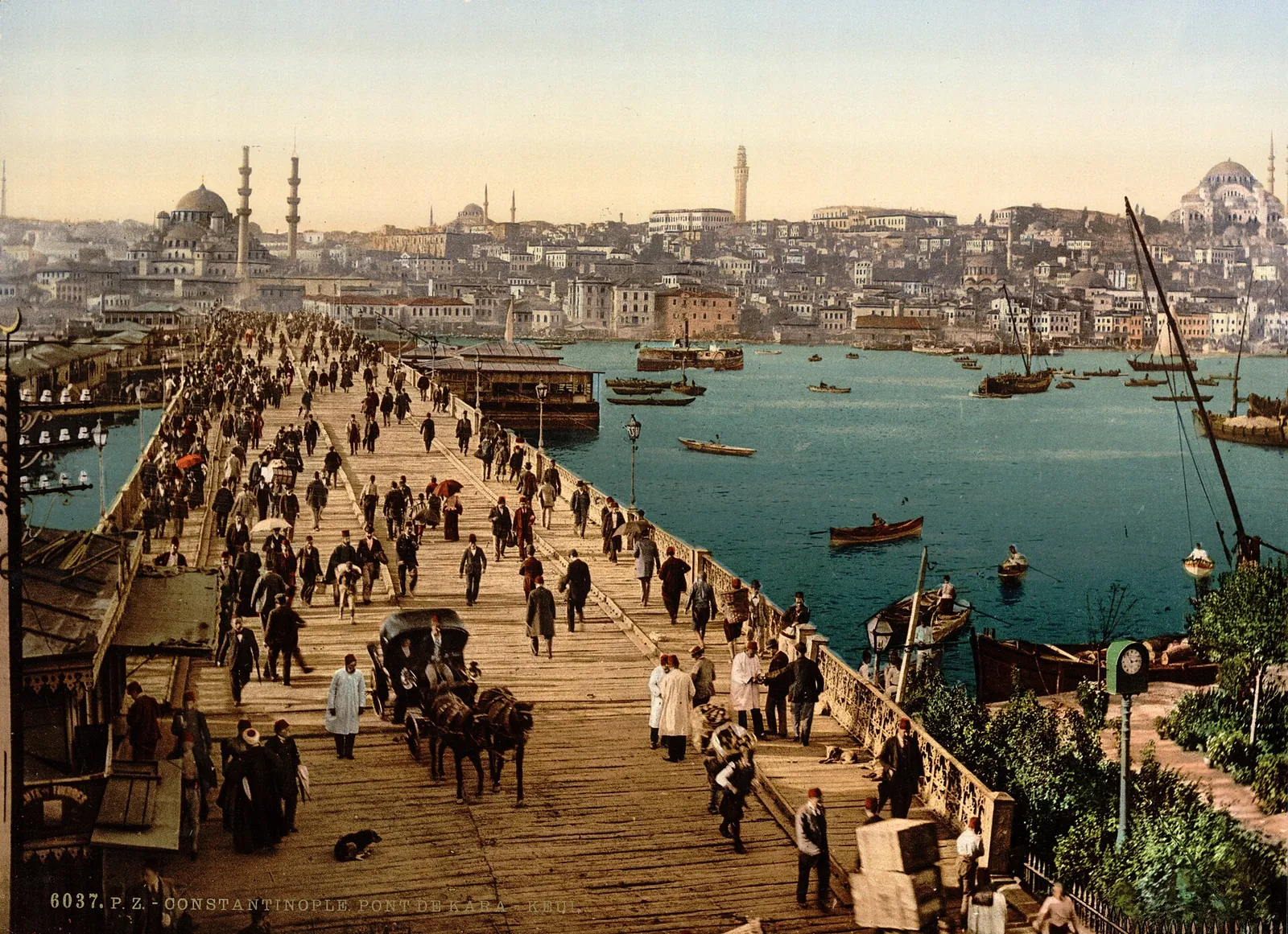 Fotografie veche colorată: Podul Galata plin de trecători, cu vapoare la chei și, pe malul celălalt, Moscheea Nouă și dealul cu Süleymaniye.