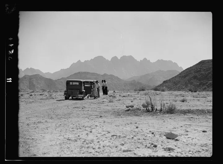 An old photograph: a wide sandy valley between bare mountains, with a clump of palm trees.