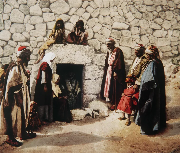 An old photograph: the low stone entrance to a tomb, with a man standing beside it.