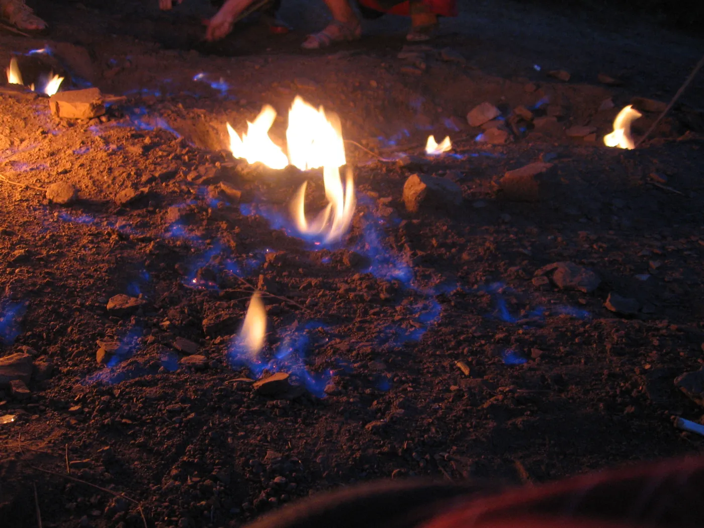 Blue and yellow flames on the seepage patch at night, with visitors' feet beside them for scale. Photo: Petr Sporer, 9 August 2018. (Photo: Wikimedia Commons, CC BY-SA 4.0)