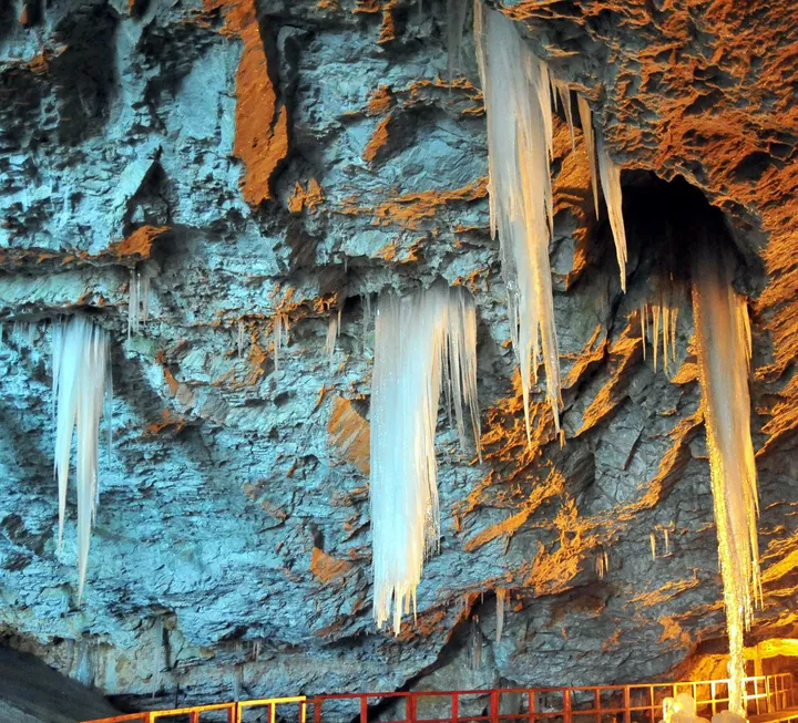 Long ice stalactites hanging from the rusty cave ceiling above the handrail