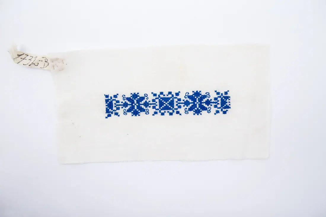 Fotografia muzeului: un model de broderie pe pânză albă, cu fir albastru, cusut de Minodora Cismaș