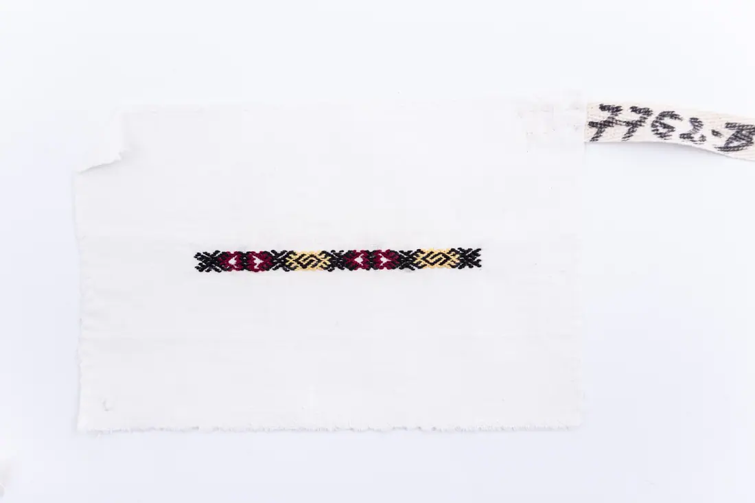 Fotografia muzeului: un model de broderie pe pânză albă, cu fir negru, galben, vișiniu, cusut de Filip Maria