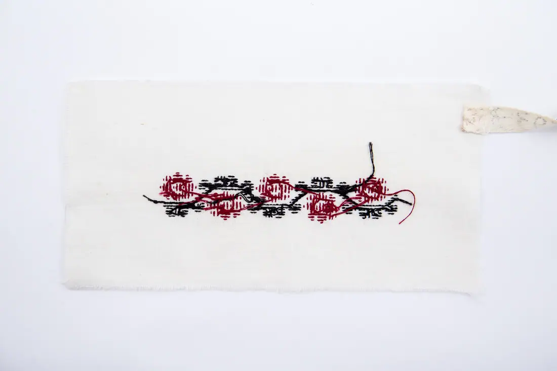 Fotografia muzeului: un model de broderie pe pânză albă, cu fir negru, roșu, cusut de Bădilă Zâna