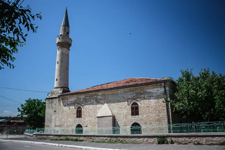 A stone mosque with a tiled roof and a white minaret, beside a paved street.