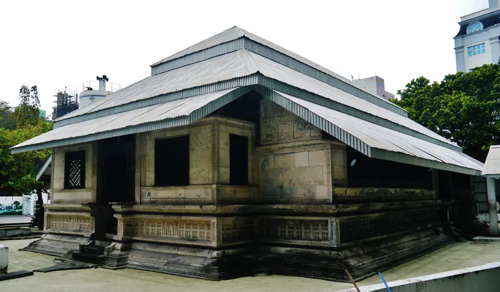 A low mosque built of coral blocks, with a stepped metal roof, among modern buildings.