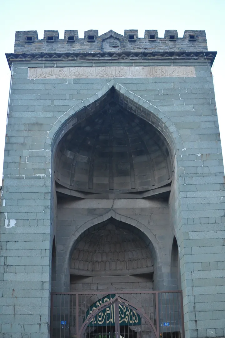 A massive grey stone gateway with a pointed arch and battlements, set in a mosque wall.
