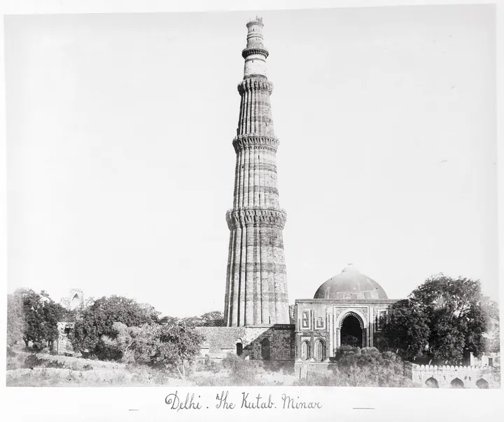 A black-and-white photograph: a fluted stone minaret tapering as it rises, far taller than the buildings around it.