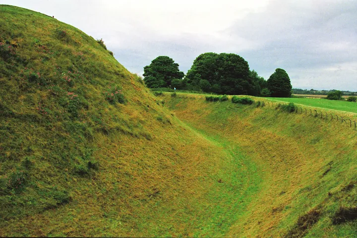 Colour photograph of a tall earthen mound ringed by a deep ditch and grass, under a cloudy sky