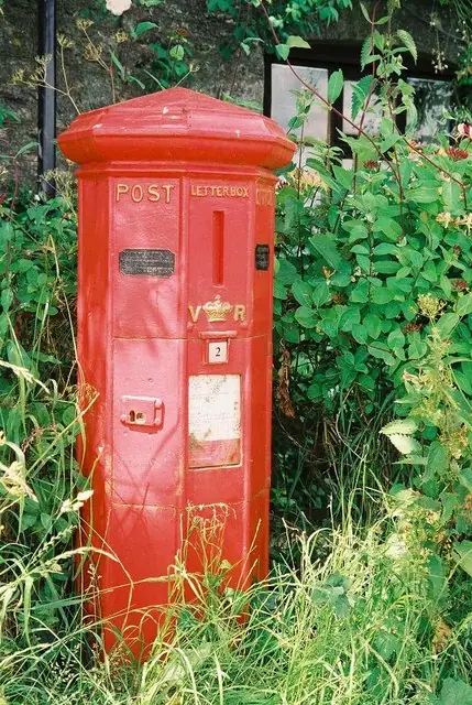 Cutia poștală din 1853 de la Barnes Cross, Holwell: o cutie roșie, poligonală, cu inscripțiile POST și LETTER BOX și cifrul VR, într-un gard viu.