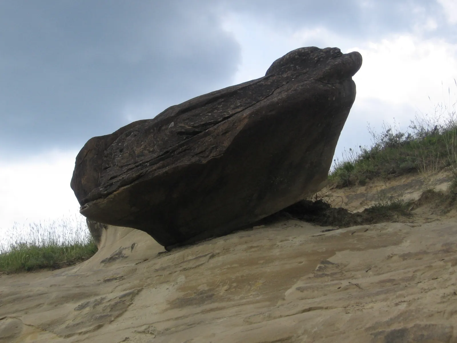 Un bolovan mare, rotunjit, de gresie închisă la culoare, stă pe un versant de nisip gălbui, sub un cer înnorat, la Ulmet