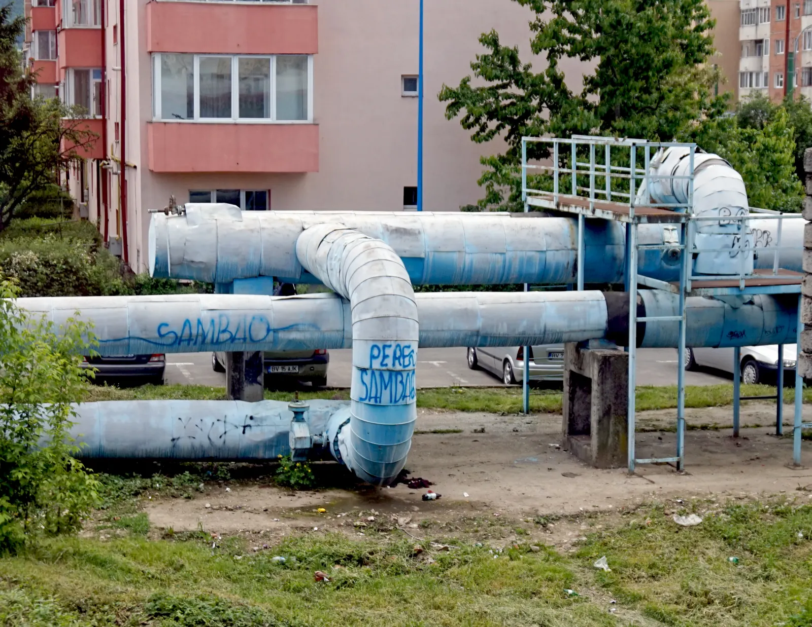 Thick district-heating pipes with peeling blue-painted insulation running above a car park between apartment blocks in Brașov