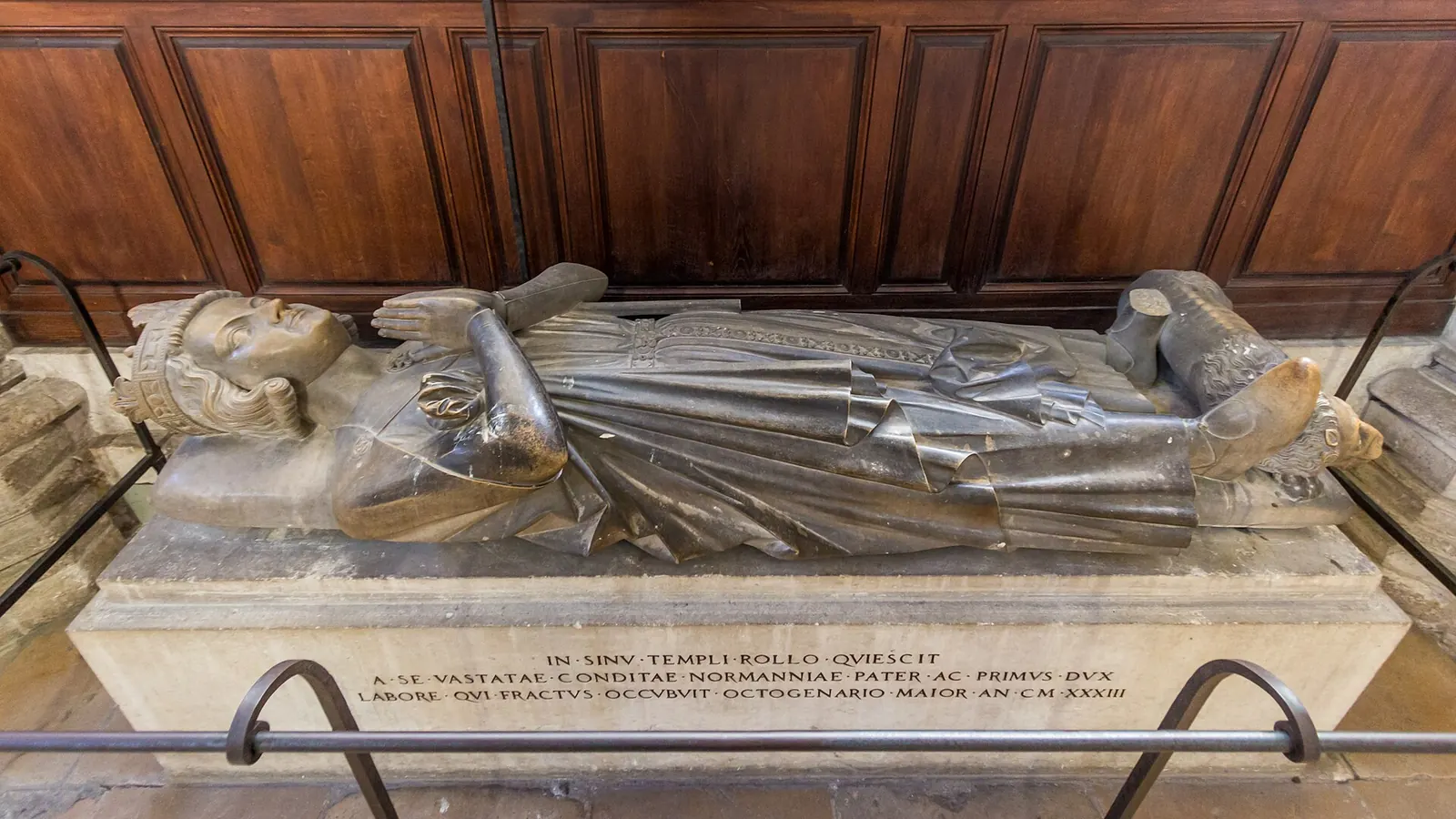 Statuie funerară de piatră a unui personaj înveșmântat, întins pe un soclu cu inscripție latină.