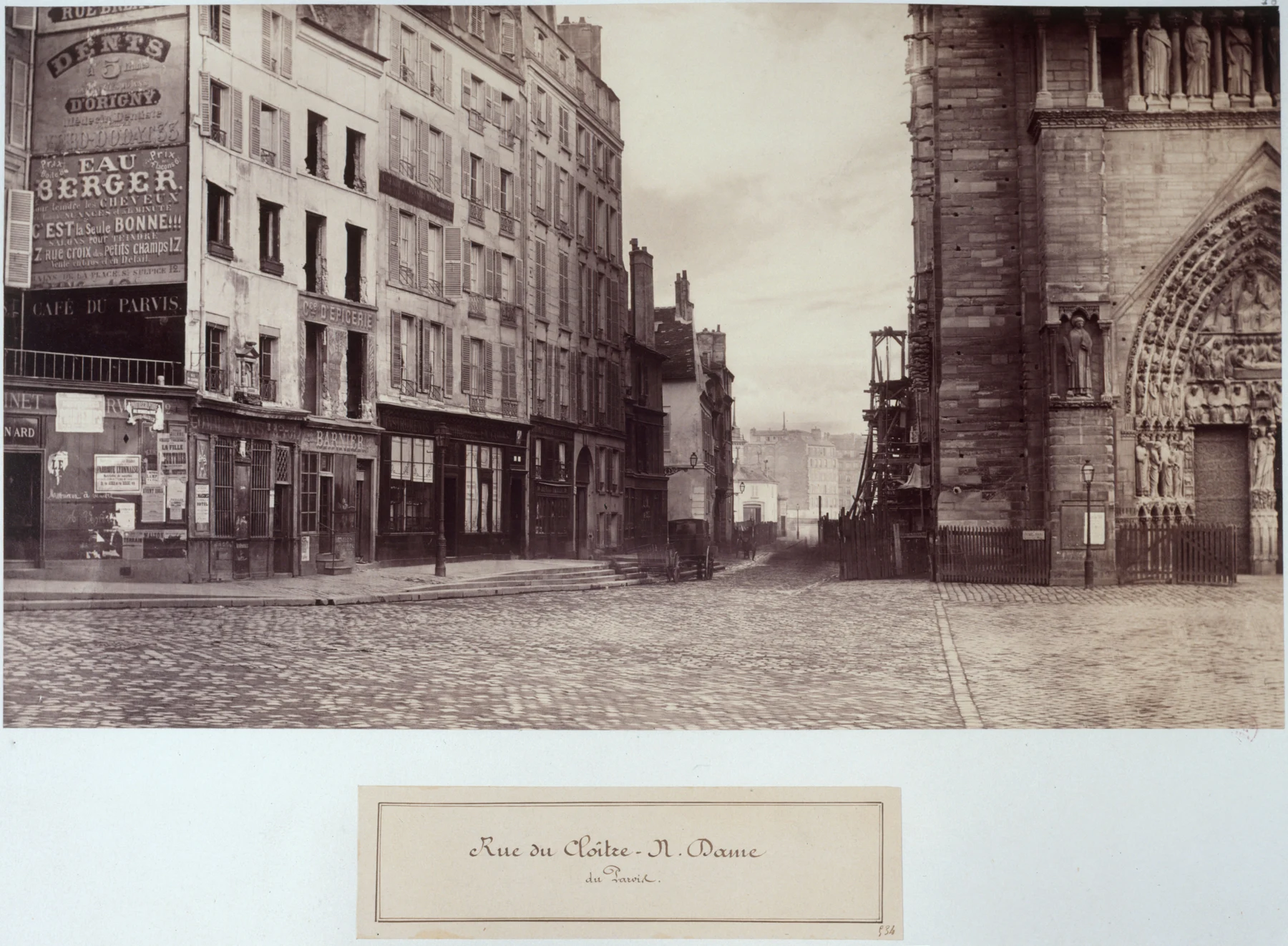 View from the parvis along a street, with houses on the left and Notre-Dame’s sculpted portal on the right.