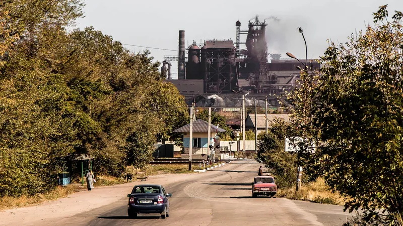 A street with cars leading to the chimneys and blast furnaces of a steelworks