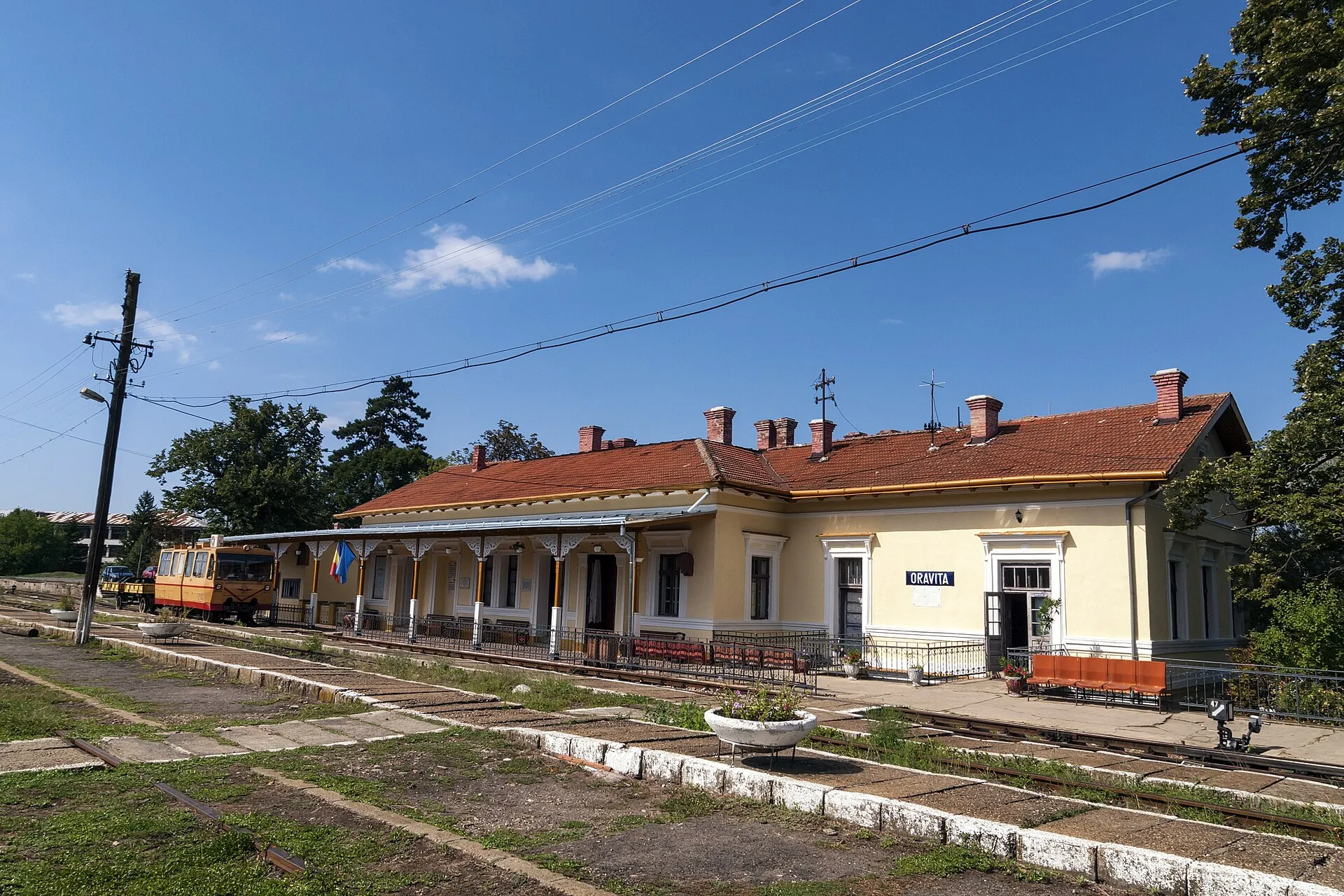 Clădirea gării Oravița, cu acoperiș roșu și arcade, văzută de pe peron.