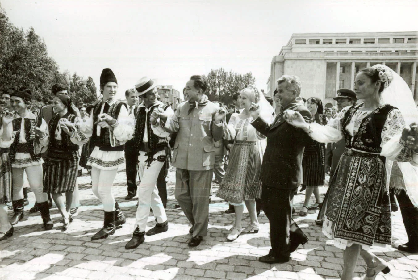Fotografie alb-negru: un șir de dansatori în costume populare românești se țin de mână pe caldarâm; printre ei, doi bărbați în haine civile, unul într-o jachetă deschisă la culoare și unul în costum închis; în dreapta se vede o clădire mare de piatră.