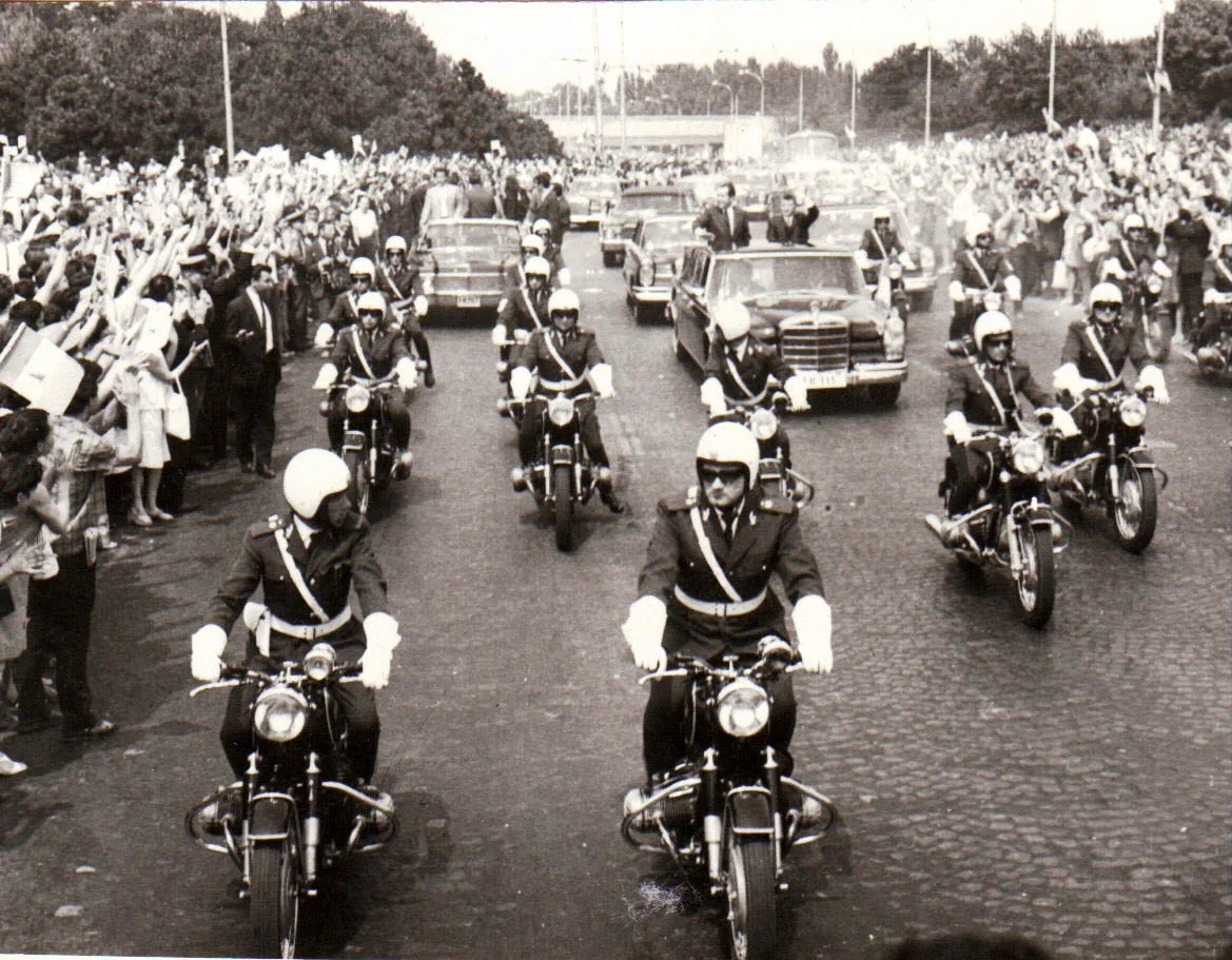 Fotografie alb-negru: motocicliști ai escortei, cu căști albe și mănuși albe, deschid coloana de limuzine pe o arteră pavată; mulțimi de oameni de ambele părți flutură mâinile și stegulețe.