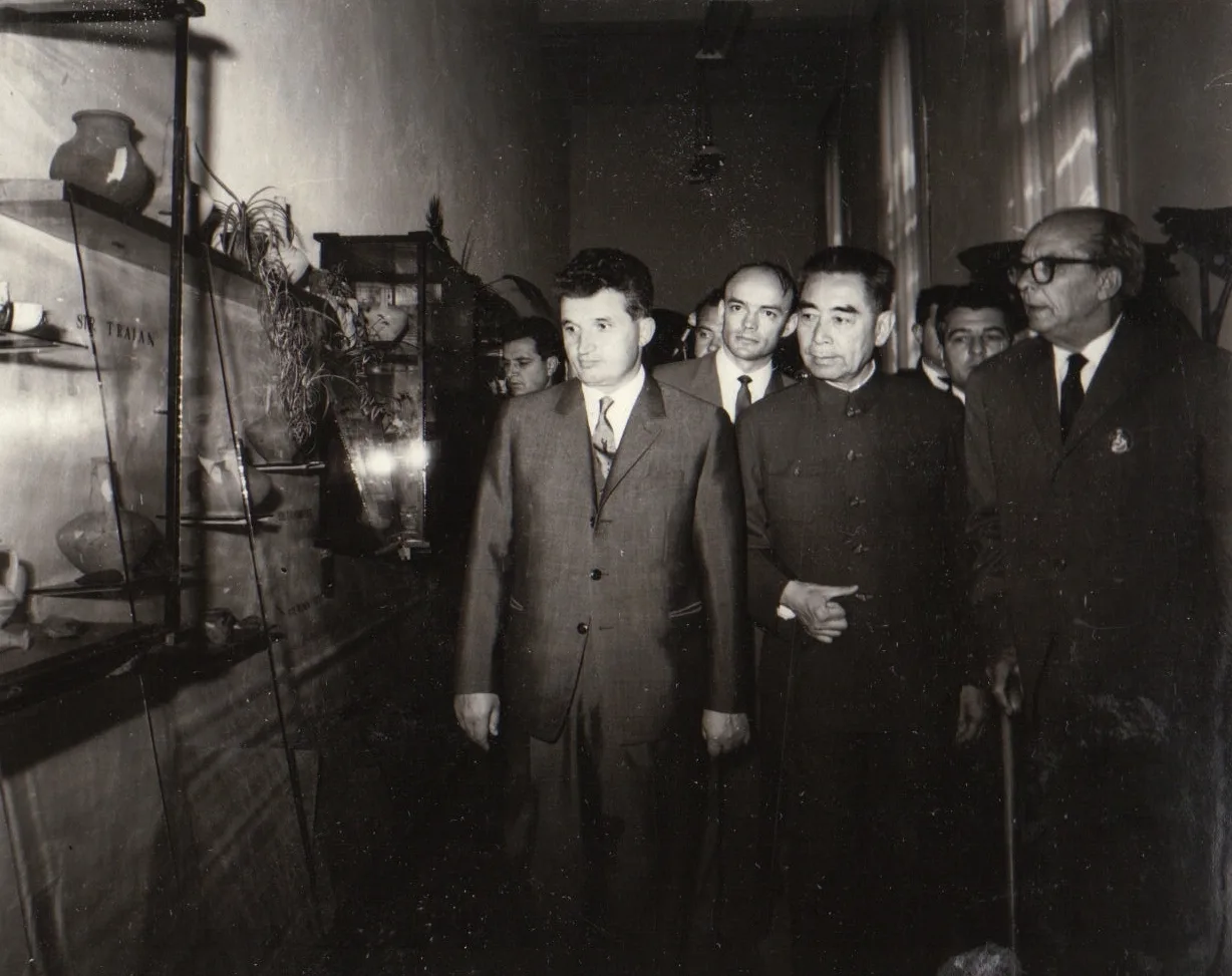 Fotografie alb-negru: într-un coridor de muzeu, lângă vitrine cu ceramică veche, merg mai mulți bărbați; în prim-plan unul în costum de stofă, lângă el unul într-o tunică închisă, iar în dreapta un bărbat în vârstă cu ochelari.