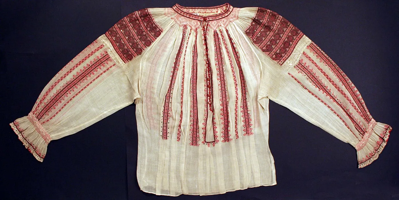 O ie albă cu broderie roșie: benzi late și întunecate pe umeri, sub ele o bandă îngustă și deschisă la culoare, apoi dungi brodate care coboară pe mâneci și pe piept.