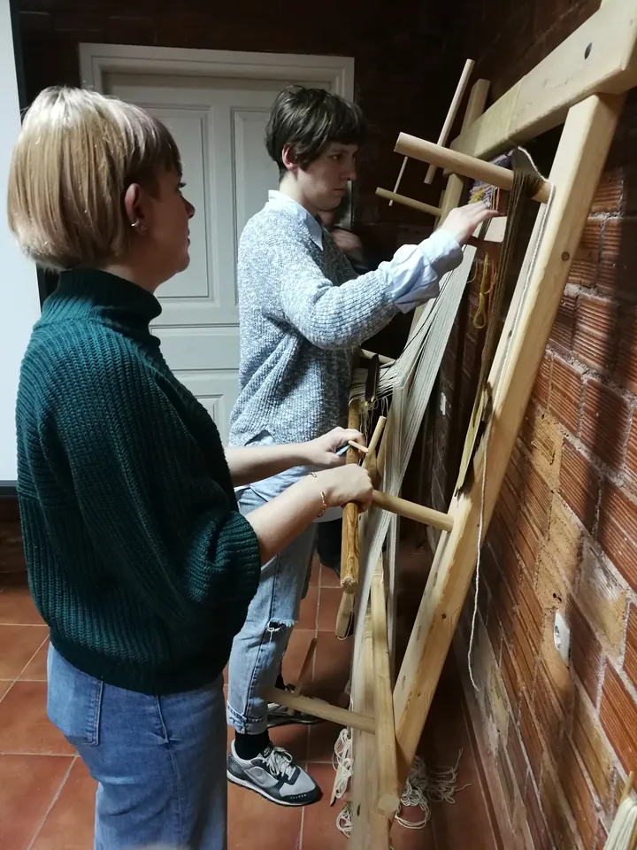 Two women working at a wooden vertical weaving loom.