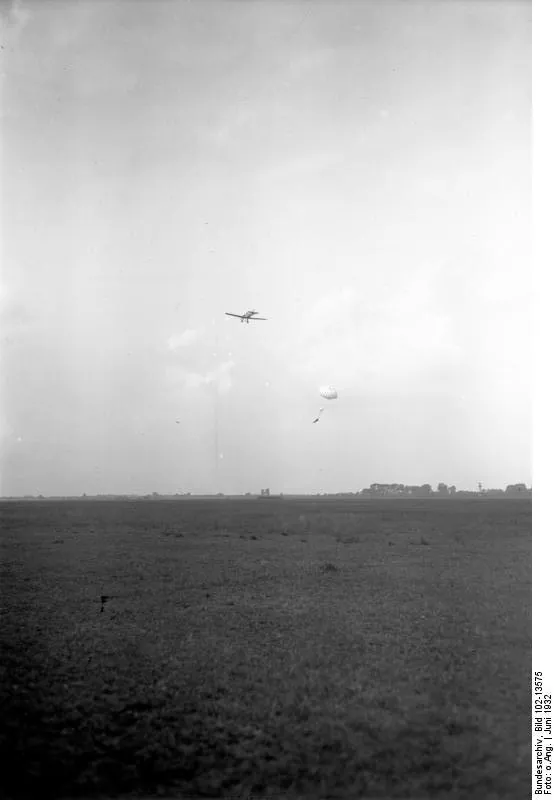 Period photograph, June 1932: a small plane with an open parachute and its jumper hanging below, over a broad field.