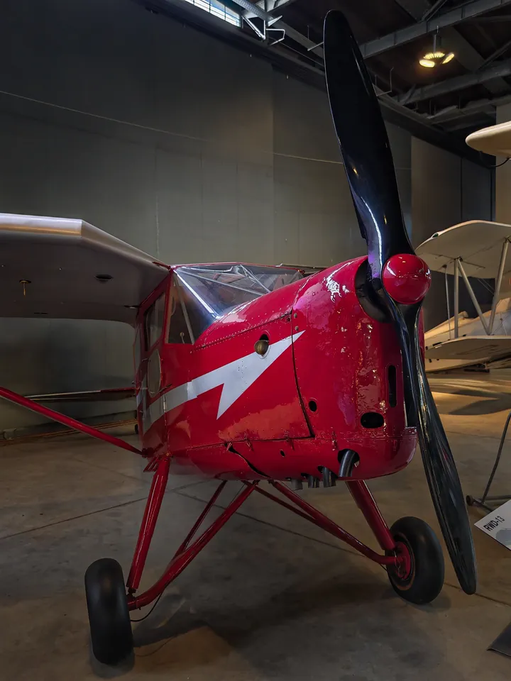 Red RWD-13 seen head-on, with a black propeller and a white lightning stripe on the nose, in a museum.