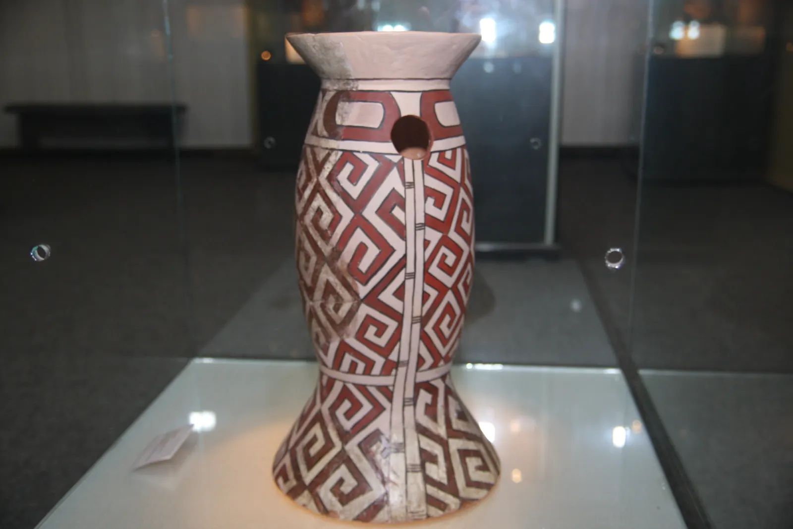 Obiect ceramic înalt, pictat cu motive geometrice roșii și negre pe fond deschis.