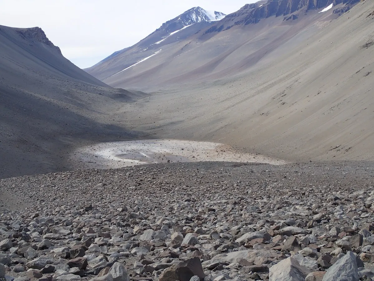 A pale, shallow salt pond on a scree-strewn valley floor between bare Antarctic mountain slopes.