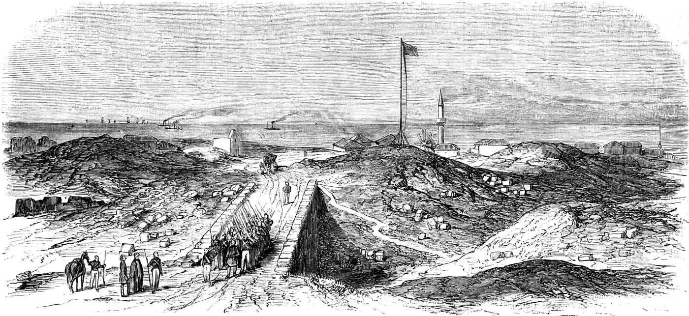 Engraving of Küstendje in 1854: soldiers march along a stone causeway between grassy ruins, with a minaret, a flagpole and low houses beyond, and steamships on the sea.