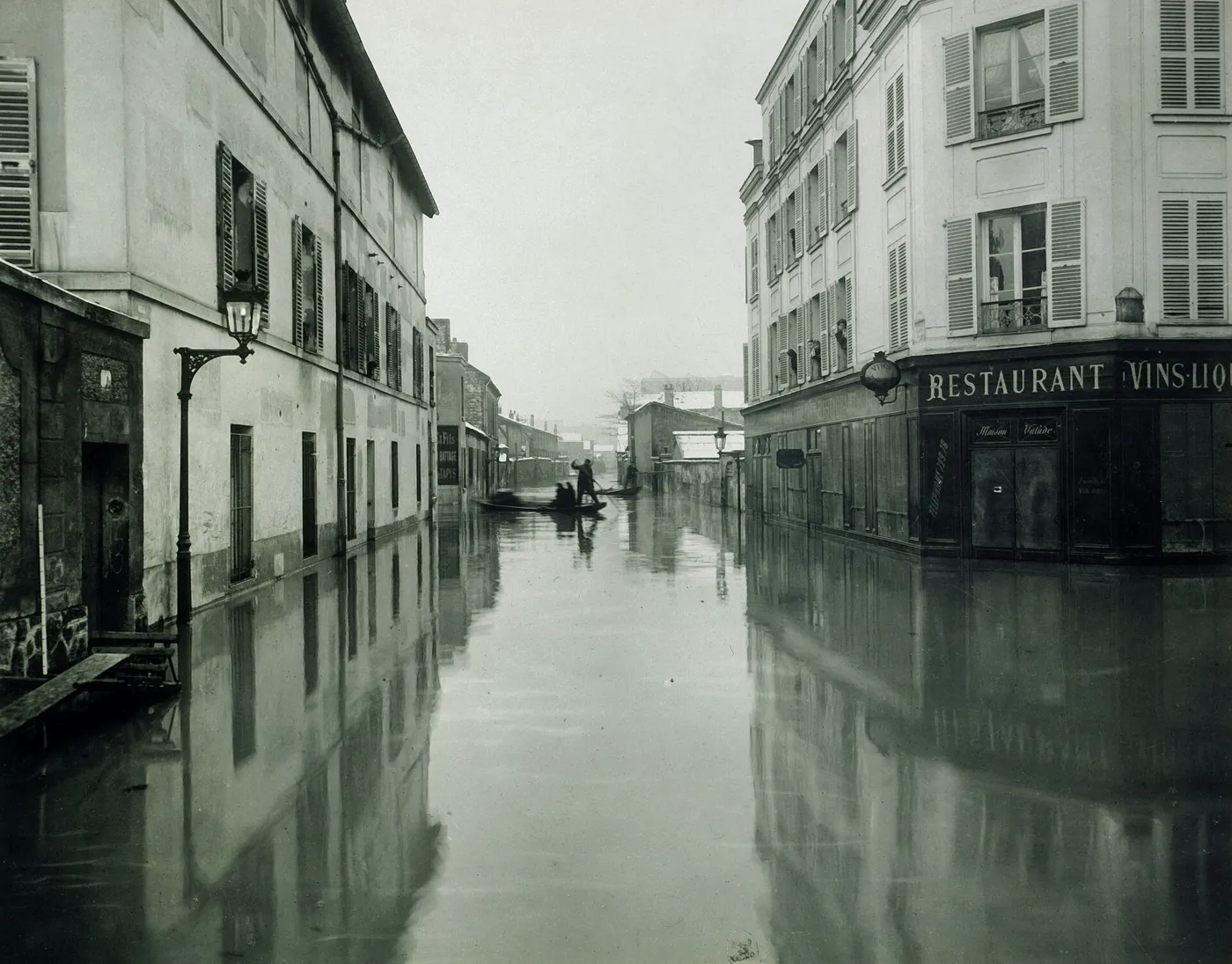 A flooded street of low houses and a corner restaurant, with a man punting a small boat in the distance and a factory gantry beyond.