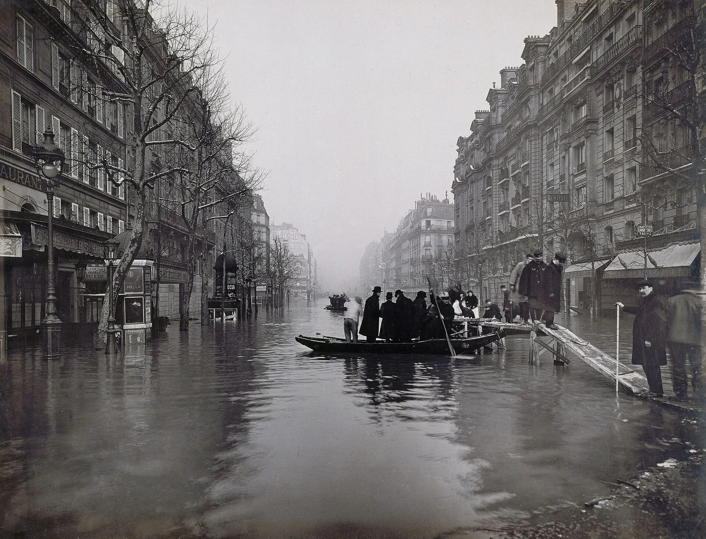 Men in hats and overcoats step from a raised wooden walkway into a rowing boat on the flooded rue de Lyon, between tall apartment buildings.