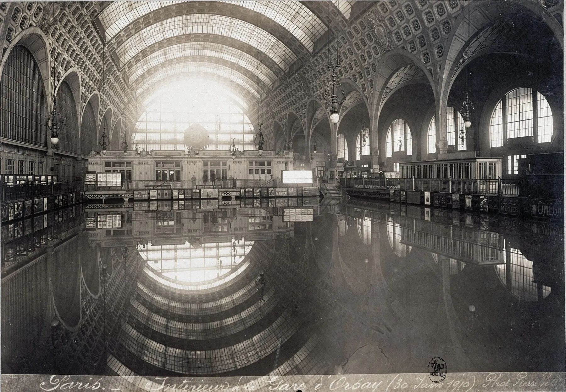 The vast iron-and-glass concourse of the Gare d'Orsay in January 1910, its floor entirely under still water that mirrors the arched glass roof.