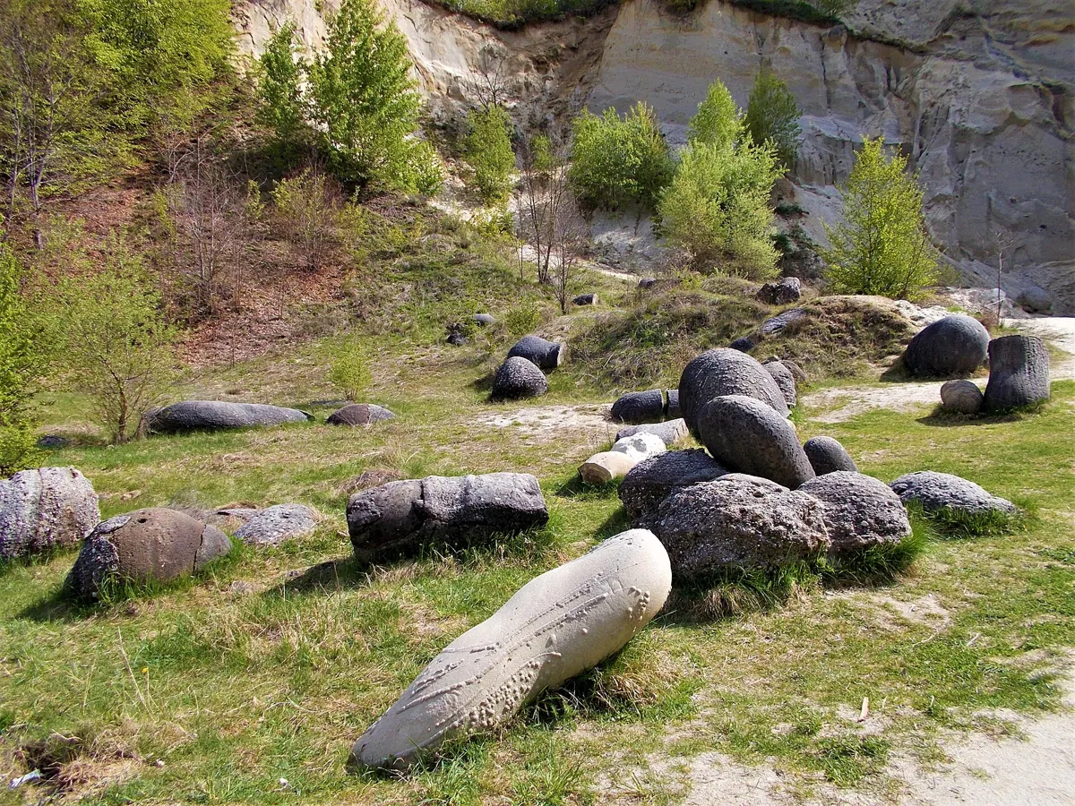 Zeci de pietre rotunjite, cenușii, împrăștiate pe iarbă, în fața unui mal abrupt de nisip.