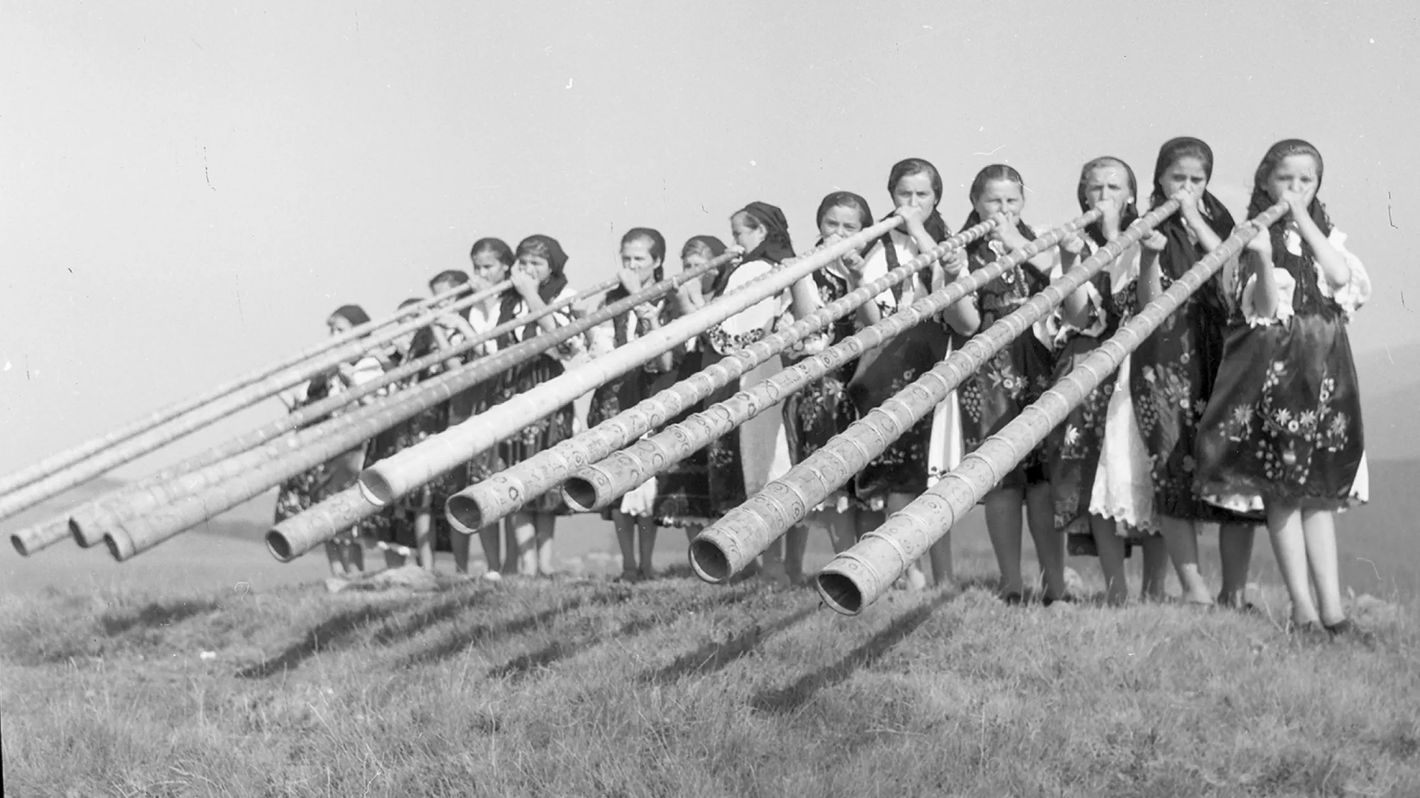 Thirteen girls in folk dress on a mountain meadow blow long tulnics, all pointing the same way.